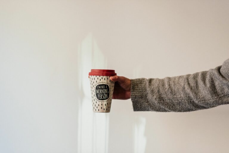 Crop anonymous person in soft wear demonstrating paper cup of takeaway hot drink with ornament and inscription